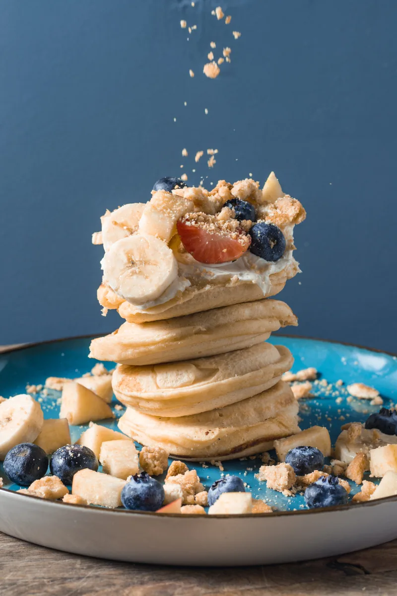 Een stapel pancakes met fruit waar poedersuiker op valt